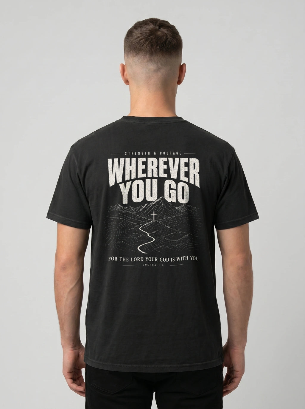 A man viewed from behind wearing a black t-shirt with a large white graphic print on the back that reads 'WHEREVER YOU GO' with a mountain and road illustration, against a light gray studio background.
