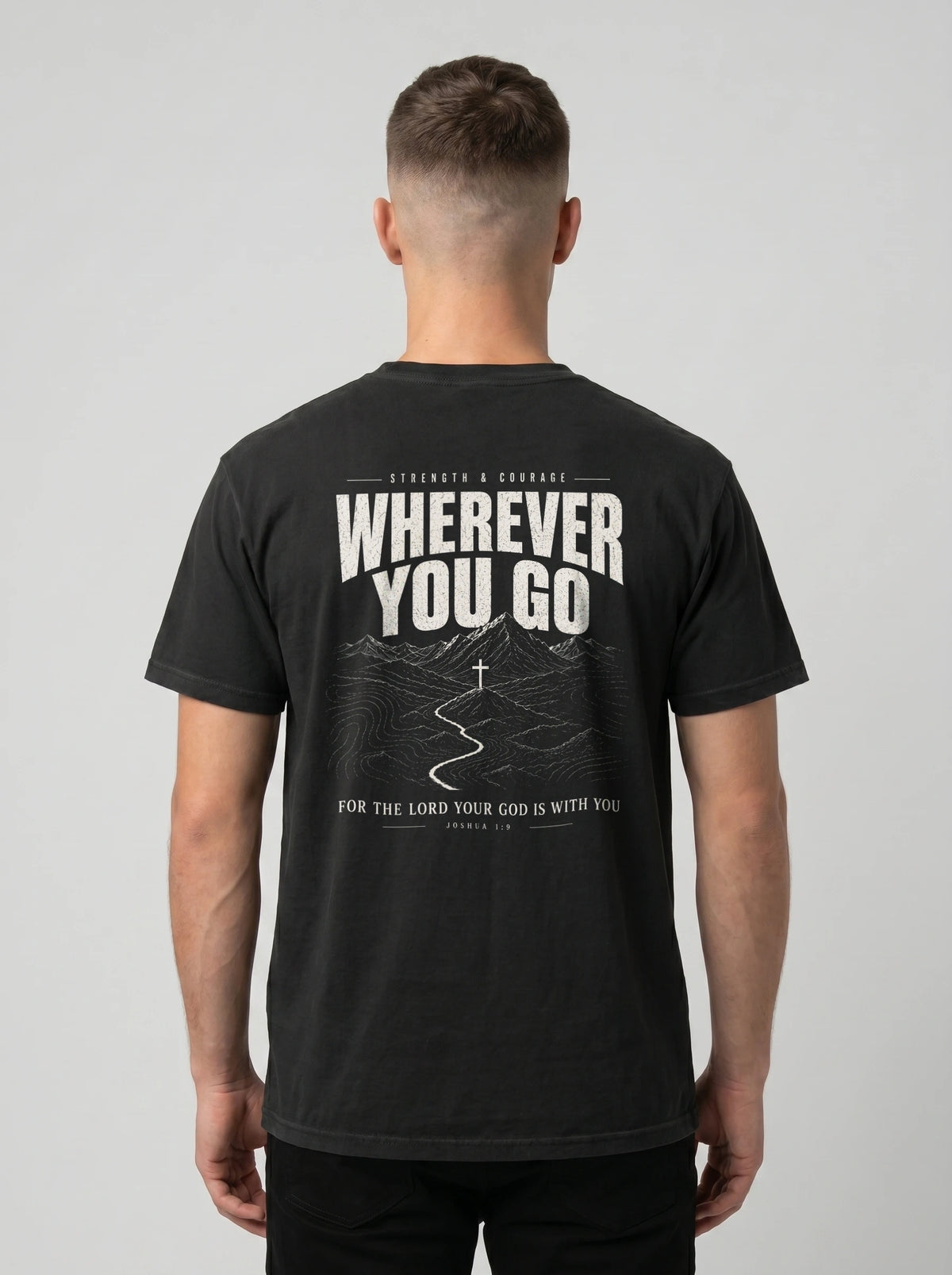 A man viewed from behind wearing a black t-shirt with a large white graphic print on the back that reads 'WHEREVER YOU GO' with a mountain and road illustration, against a light gray studio background.