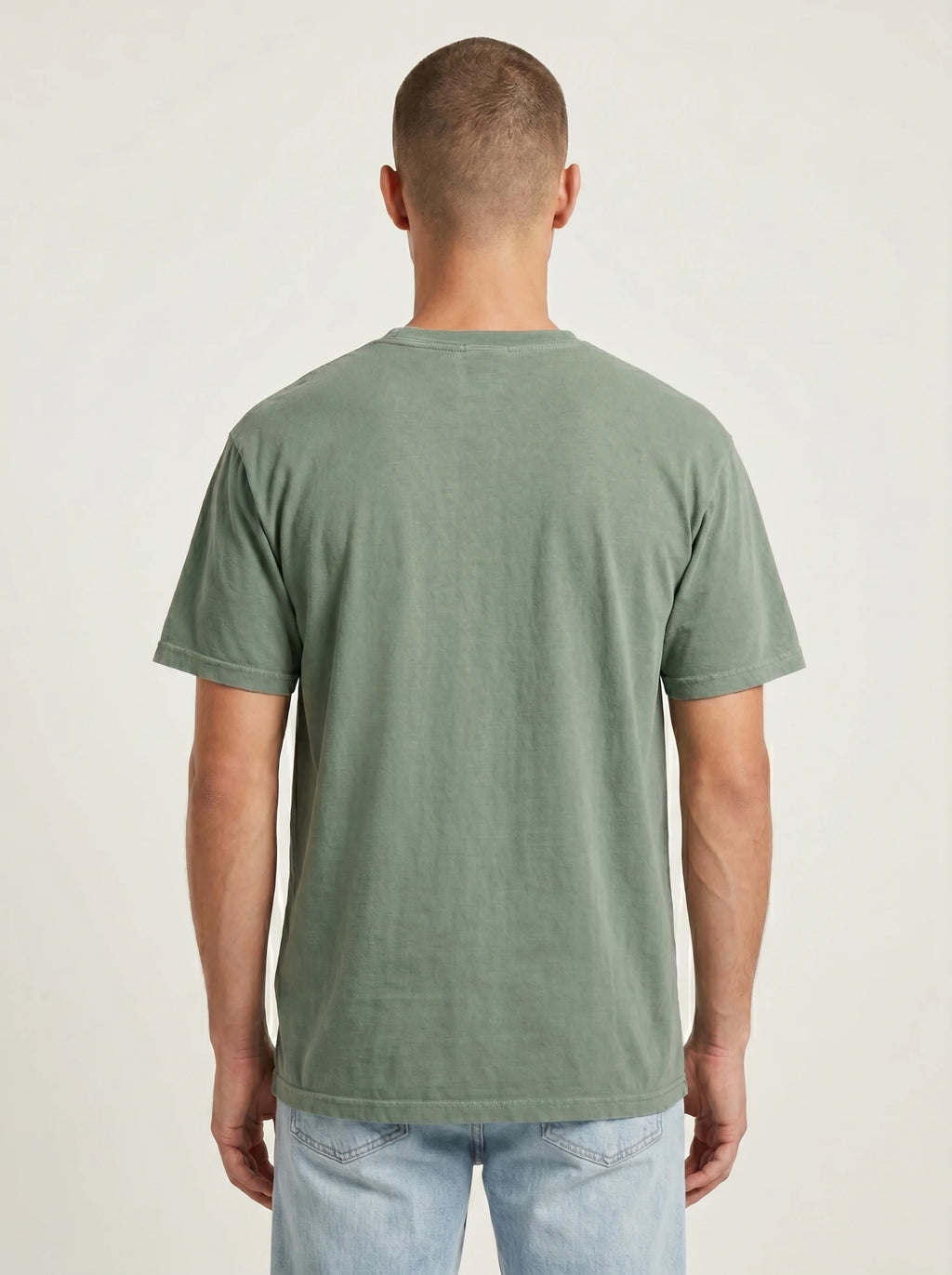A man viewed from behind wearing a muted sage green short-sleeve t-shirt and light blue jeans, standing against a plain off-white studio background.