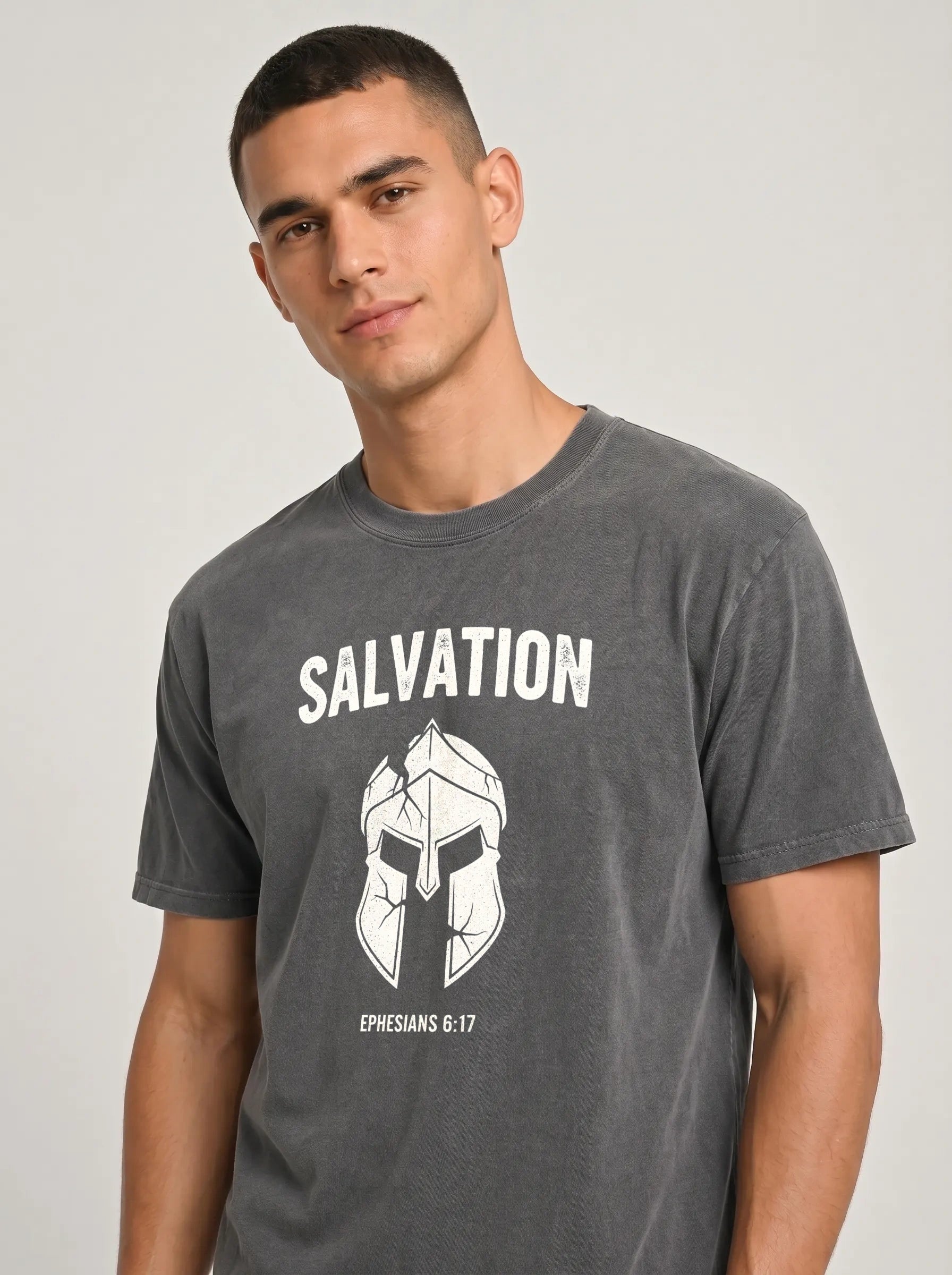 A man wearing a dark gray crewneck t-shirt with a white graphic print of a Spartan helmet and the word 'SALVATION' on the chest, set against a plain light gray background.