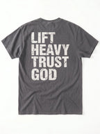 A dark gray crewneck t-shirt shown from the back, featuring the phrase 'LIFT HEAVY TRUST GOD' printed in large, distressed white block letters.