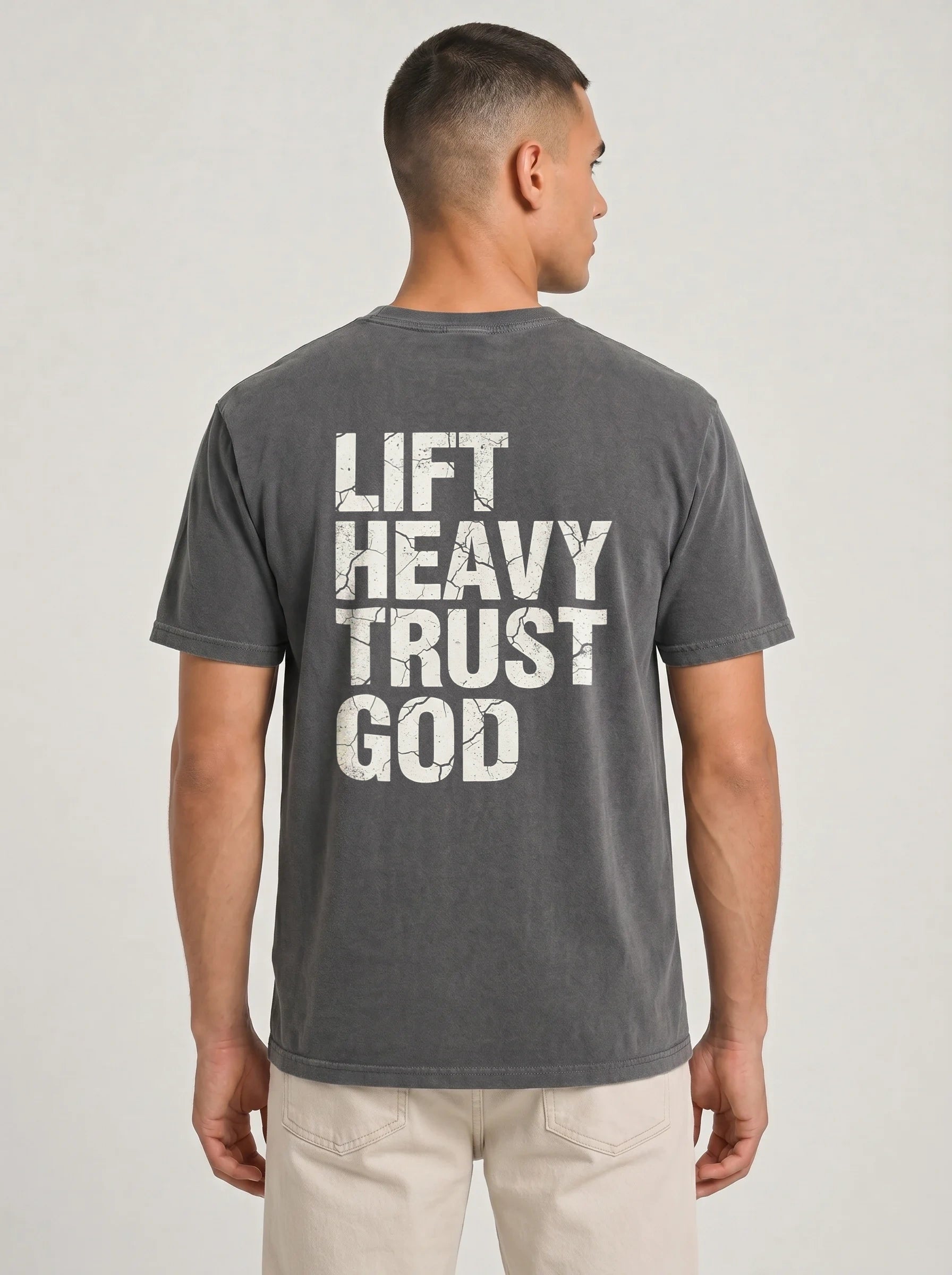 A man wearing a dark gray t-shirt with distressed white text reading 'LIFT HEAVY TRUST GOD' on the back, shown from a rear three-quarter view against a light gray studio background.