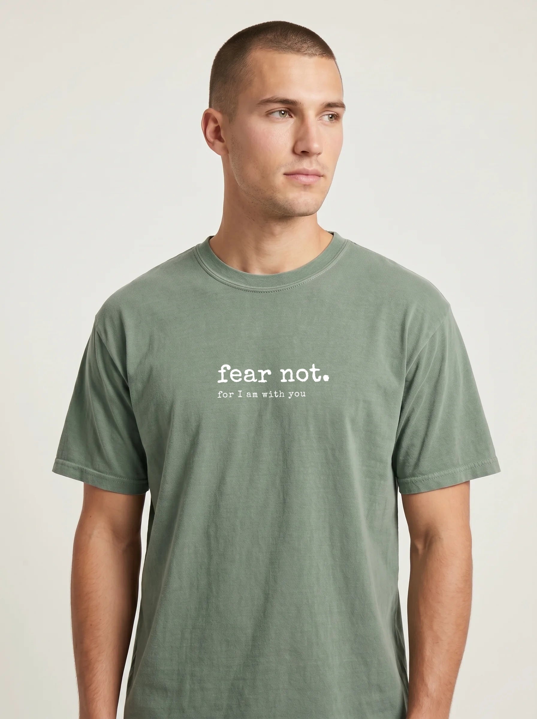 A man wearing a muted sage green crewneck t-shirt with the white printed text 'fear not. For I am with you' on the chest, photographed against a neutral light gray background.