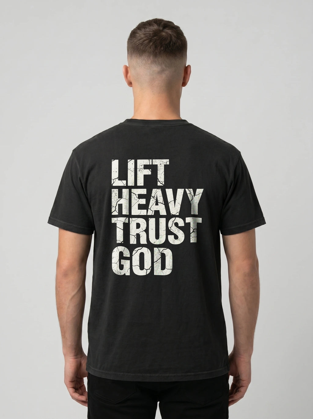 A man viewed from behind wearing a black t-shirt with the words 'LIFT HEAVY TRUST GOD' printed in distressed white block letters on the back, set against a plain light gray background.