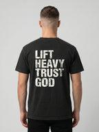 A man viewed from behind wearing a black t-shirt with the words 'LIFT HEAVY TRUST GOD' printed in distressed white block letters on the back, set against a plain light gray background.