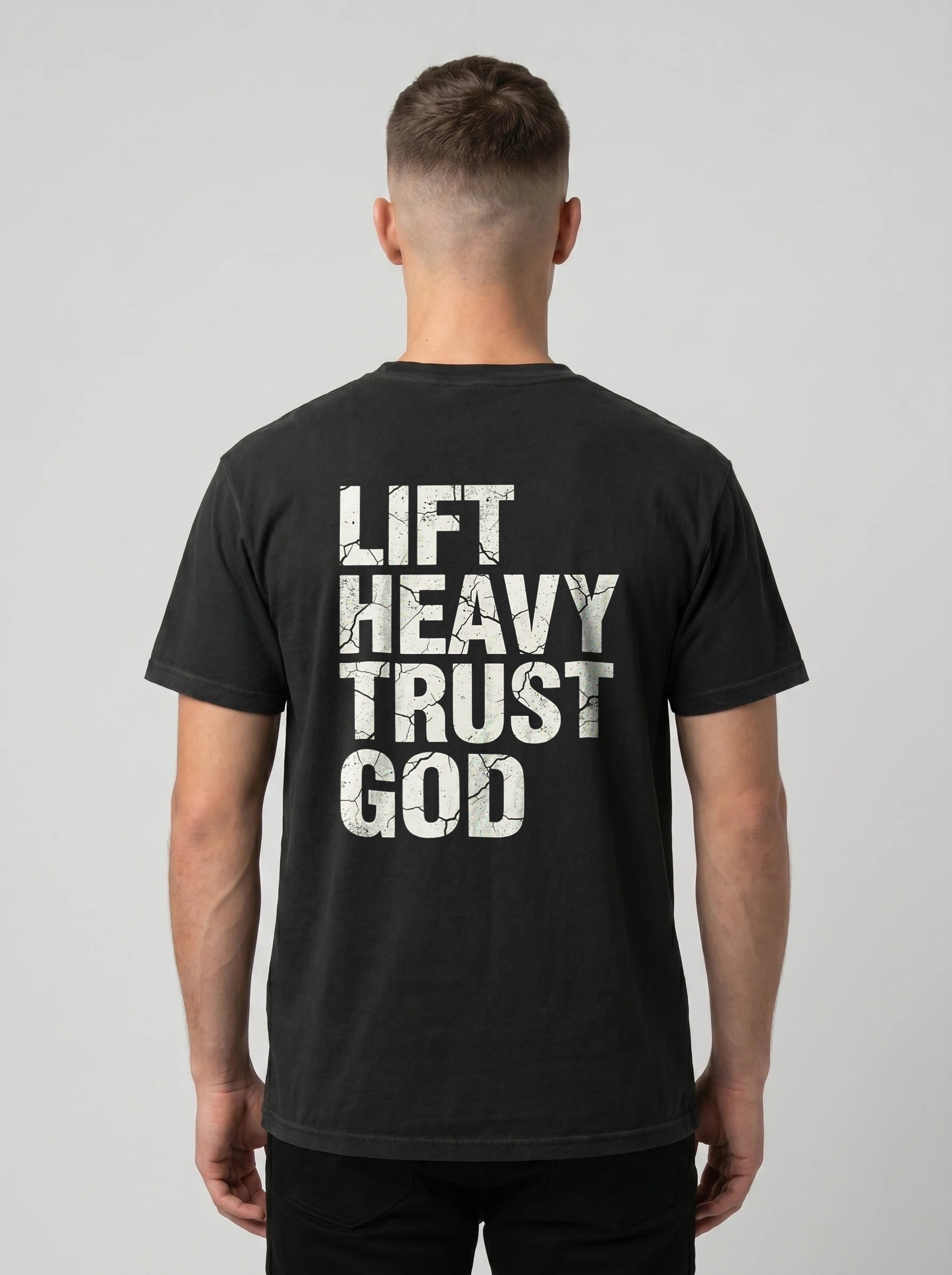 A man viewed from behind wearing a black t-shirt with the words 'LIFT HEAVY TRUST GOD' printed in distressed white block letters on the back, set against a plain light gray background.