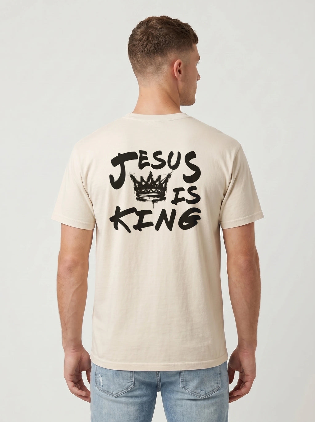 A man viewed from the back wearing a light beige t-shirt with a black graphic print that reads 'JESUS IS KING' with a crown illustration, paired with light blue jeans against a plain light gray background.