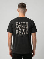 A man viewed from behind wearing a black t-shirt with a large distressed white graphic on the back that reads 'FAITH OVER FEAR' with smaller text underneath, set against a plain light gray background.