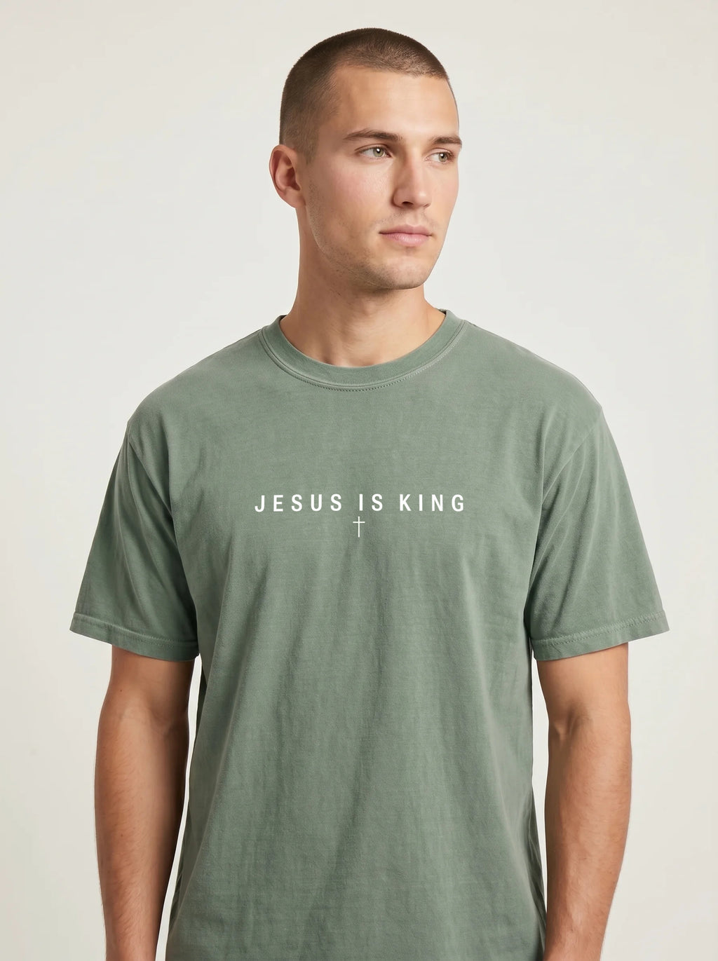 A man wearing a muted sage green crewneck t-shirt with the white printed text 'JESUS IS KING' and a small cross on the chest, photographed against a light gray studio background.