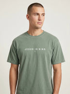 A man wearing a muted sage green crewneck t-shirt with the white printed text 'JESUS IS KING' and a small cross on the chest, photographed against a light gray studio background.