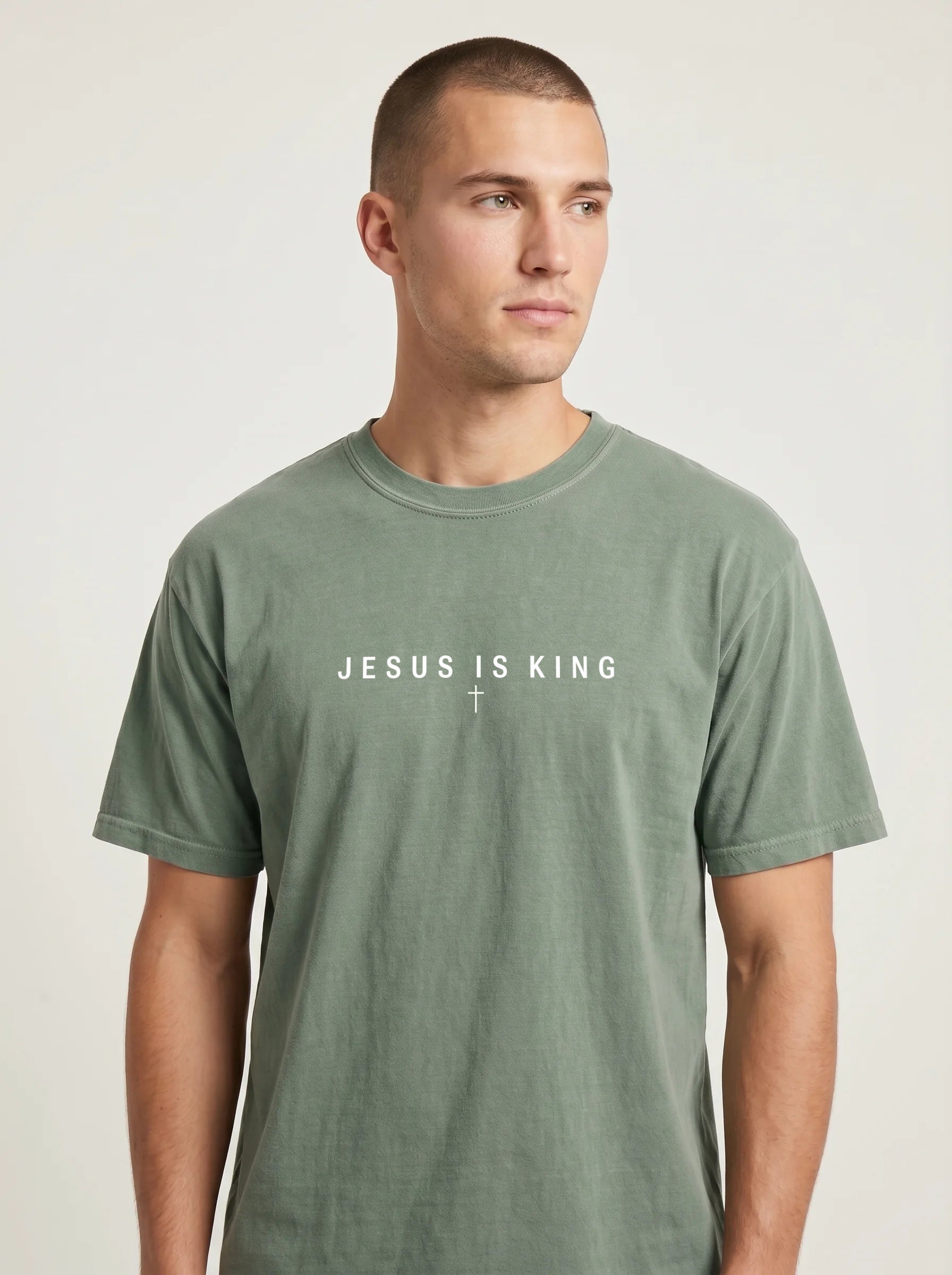 A man wearing a muted sage green crewneck t-shirt with the white printed text 'JESUS IS KING' and a small cross on the chest, photographed against a light gray studio background.