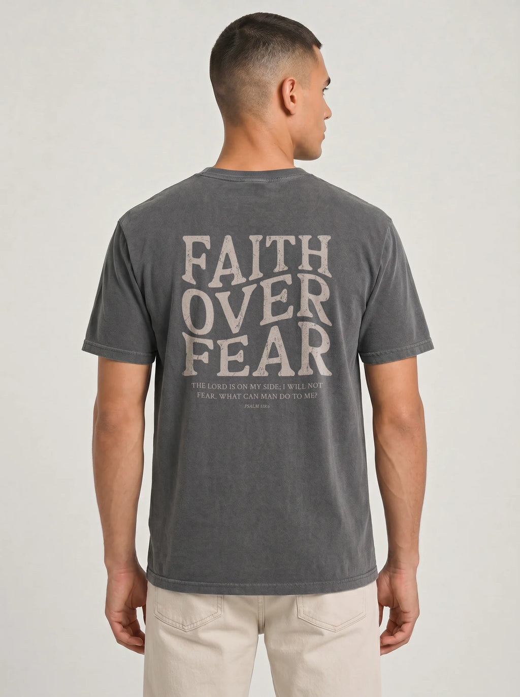 A man viewed from the back wearing a dark gray graphic t-shirt with 'FAITH OVER FEAR' printed in a distressed cream font, paired with light beige pants, against a light gray studio background.