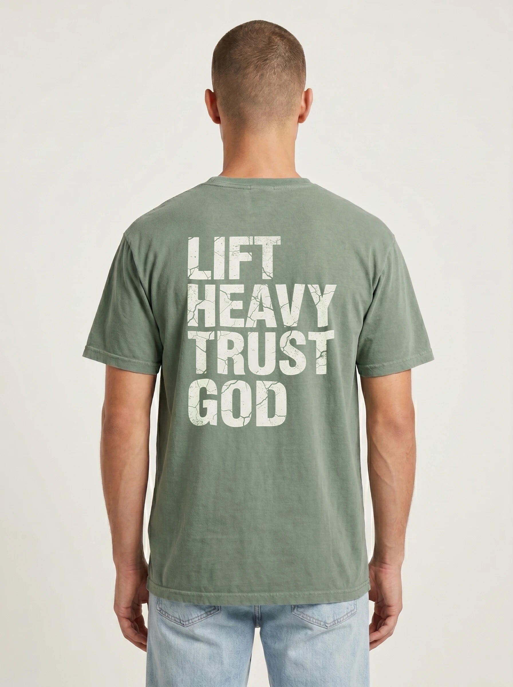A man viewed from behind wearing a sage green t-shirt with the distressed white text 'LIFT HEAVY TRUST GOD' printed on the back, paired with light blue jeans against a light gray studio background.
