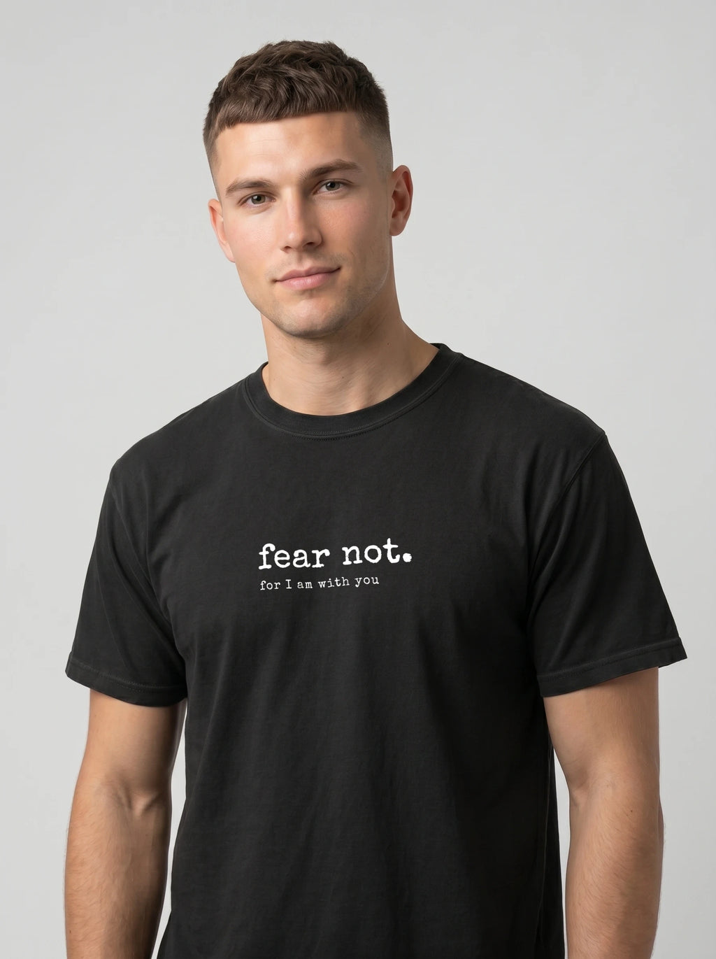 A man wearing a black crewneck t-shirt with the white printed text 'fear not. for I am with you' on the chest, photographed against a solid light gray background.