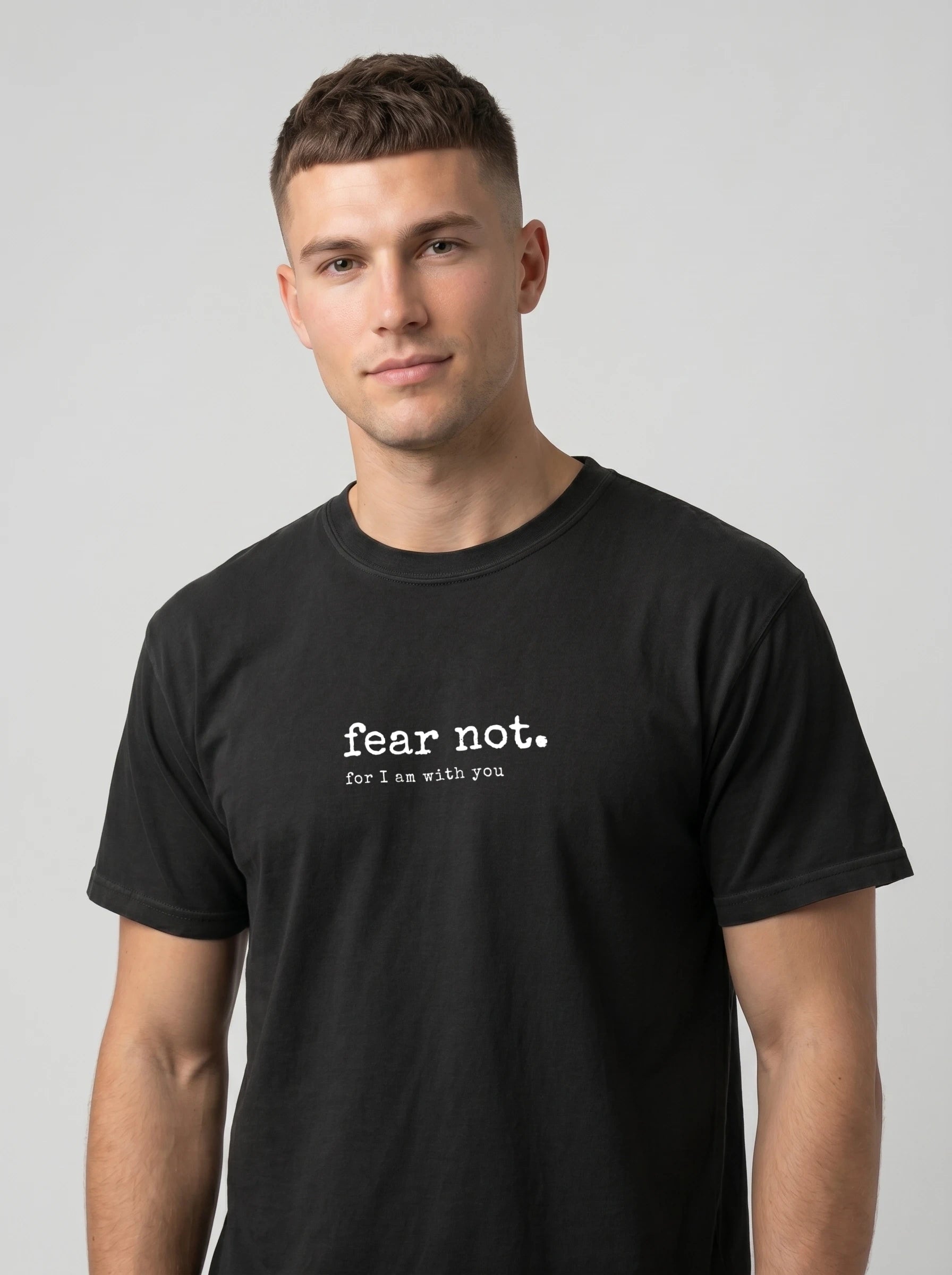 A man wearing a black crewneck t-shirt with the white printed text 'fear not. for I am with you' on the chest, photographed against a solid light gray background.