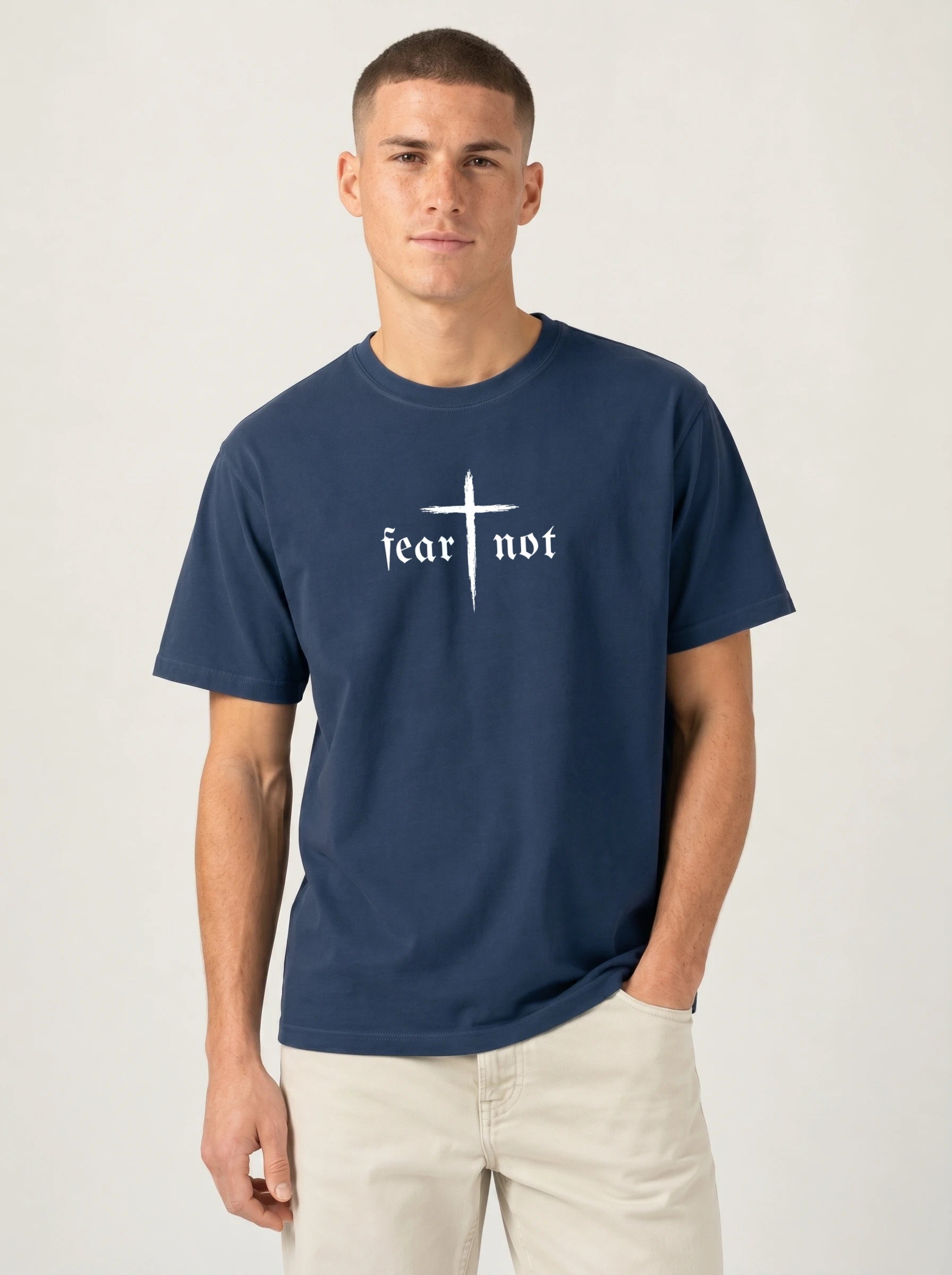 A man wearing a navy blue t-shirt with a white graphic print that reads 'fear not' under a cross, paired with light beige pants, against a plain light gray background.