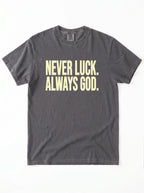 Faded charcoal T-shirt laid flat with pale yellow text reading “Never luck. Always God.”