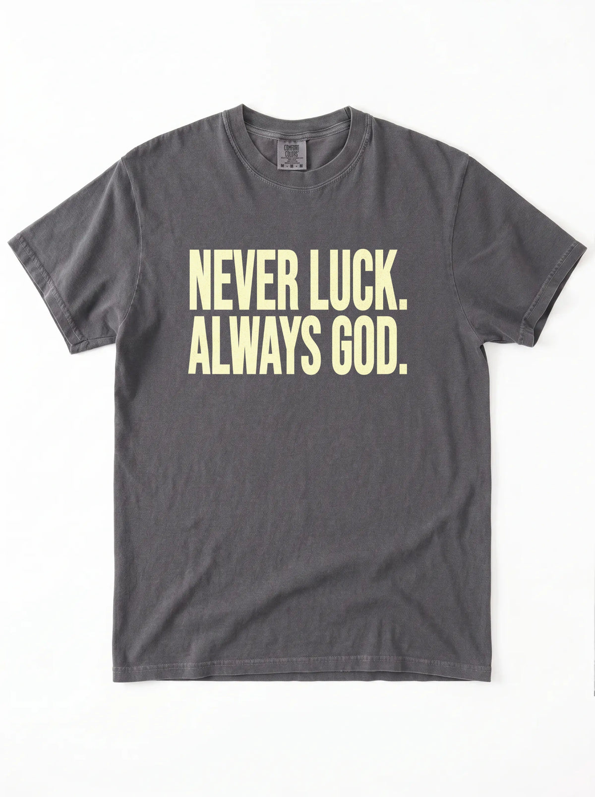 Faded charcoal T-shirt laid flat with pale yellow text reading “Never luck. Always God.”