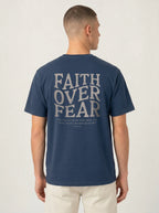 A man viewed from the back wearing a navy blue t-shirt with a large light gray graphic text reading 'FAITH OVER FEAR' and a biblical quote underneath, paired with off-white pants, against a plain light gray background.