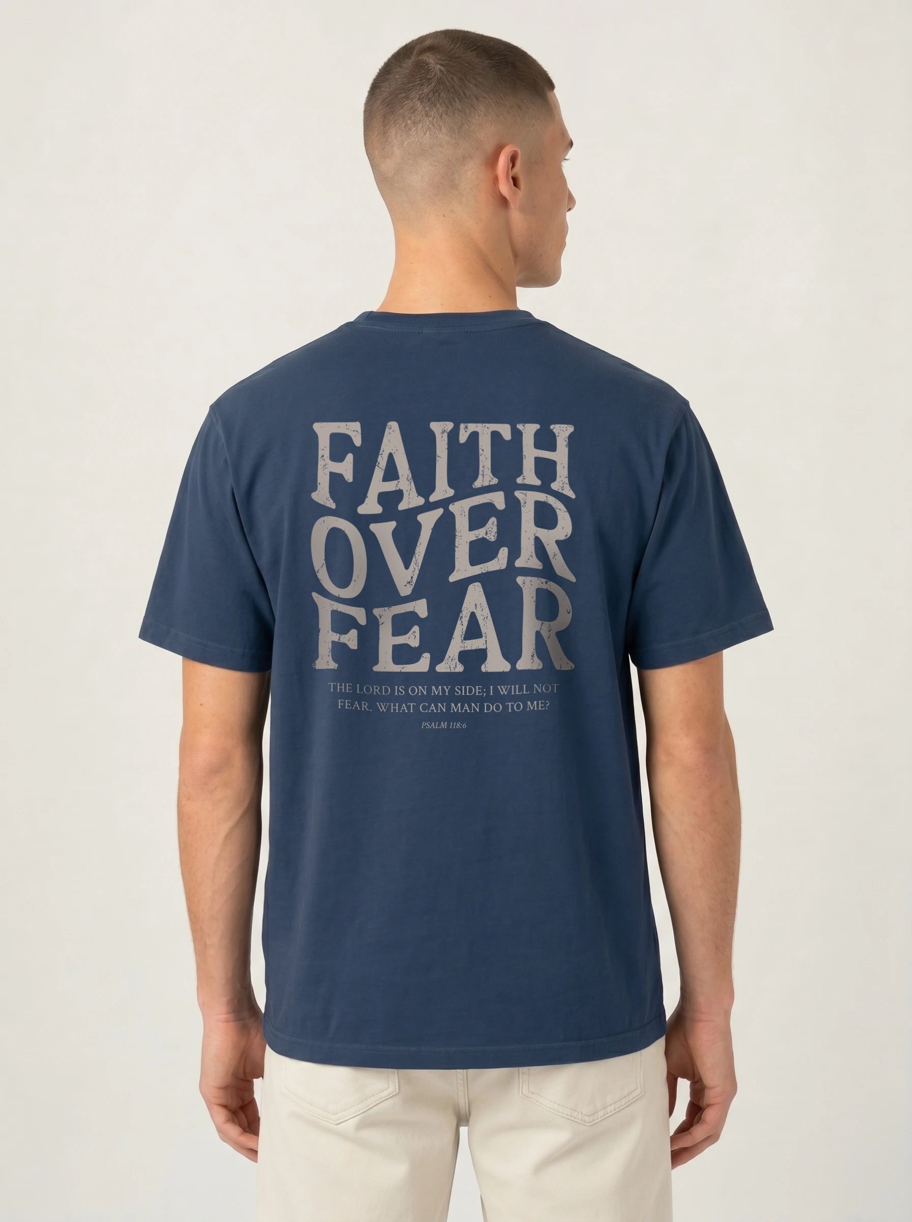 A man viewed from the back wearing a navy blue t-shirt with a large light gray graphic text reading 'FAITH OVER FEAR' and a biblical quote underneath, paired with off-white pants, against a plain light gray background.