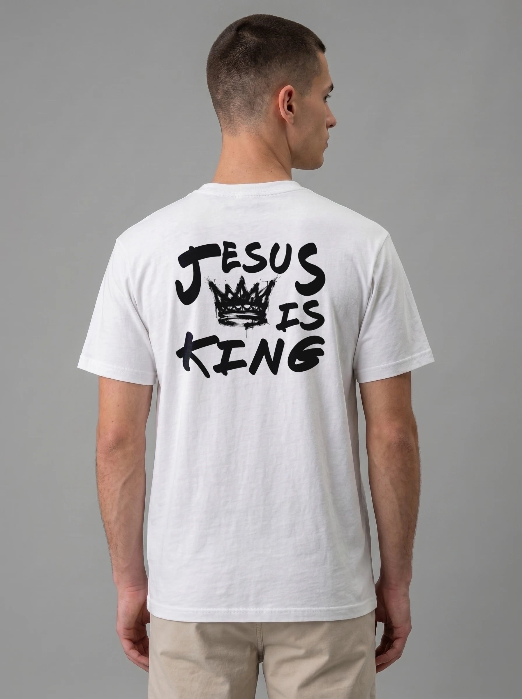 A man viewed from the back wearing a white t-shirt with a black graphic print that reads 'JESUS IS KING' with a crown illustration, paired with khaki pants against a solid gray studio background.