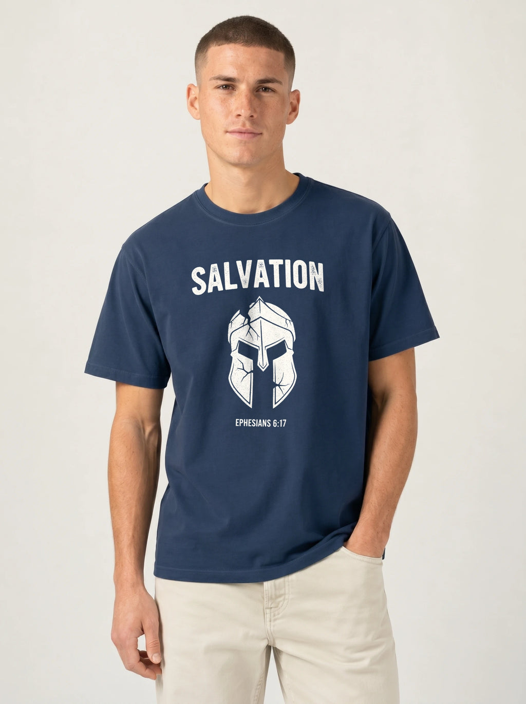A man wearing a navy blue t-shirt with a white graphic print of a helmet and the word 'SALVATION' on the front, paired with light beige pants, against a plain light gray background.