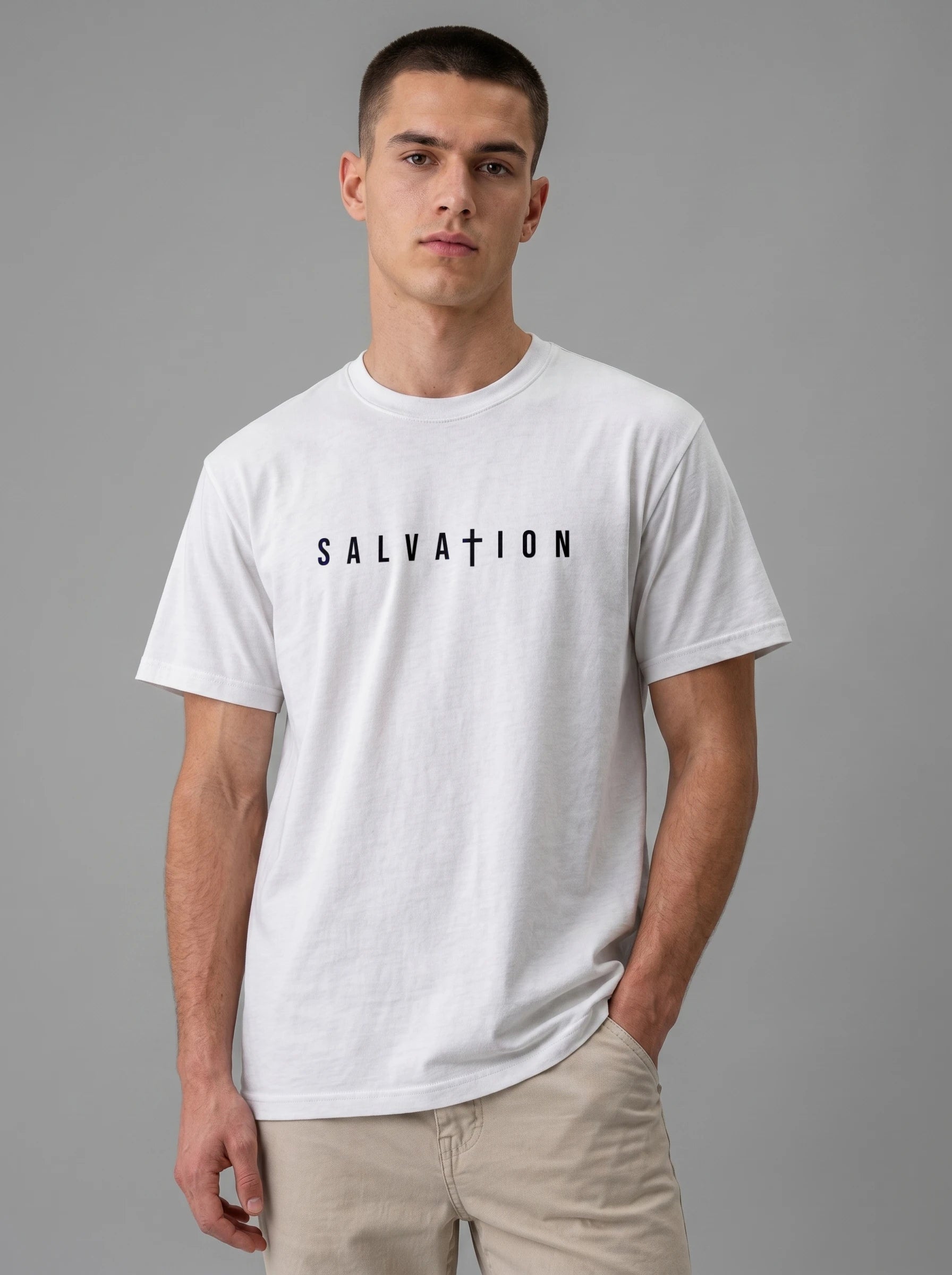 A man wearing a white crewneck t-shirt with the word 'SALVATION' printed in black across the chest, paired with beige pants, against a solid gray studio background.