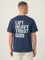 A man viewed from the back wearing a navy blue t-shirt with a distressed white graphic text that reads 'LIFT HEAVY TRUST GOD'.