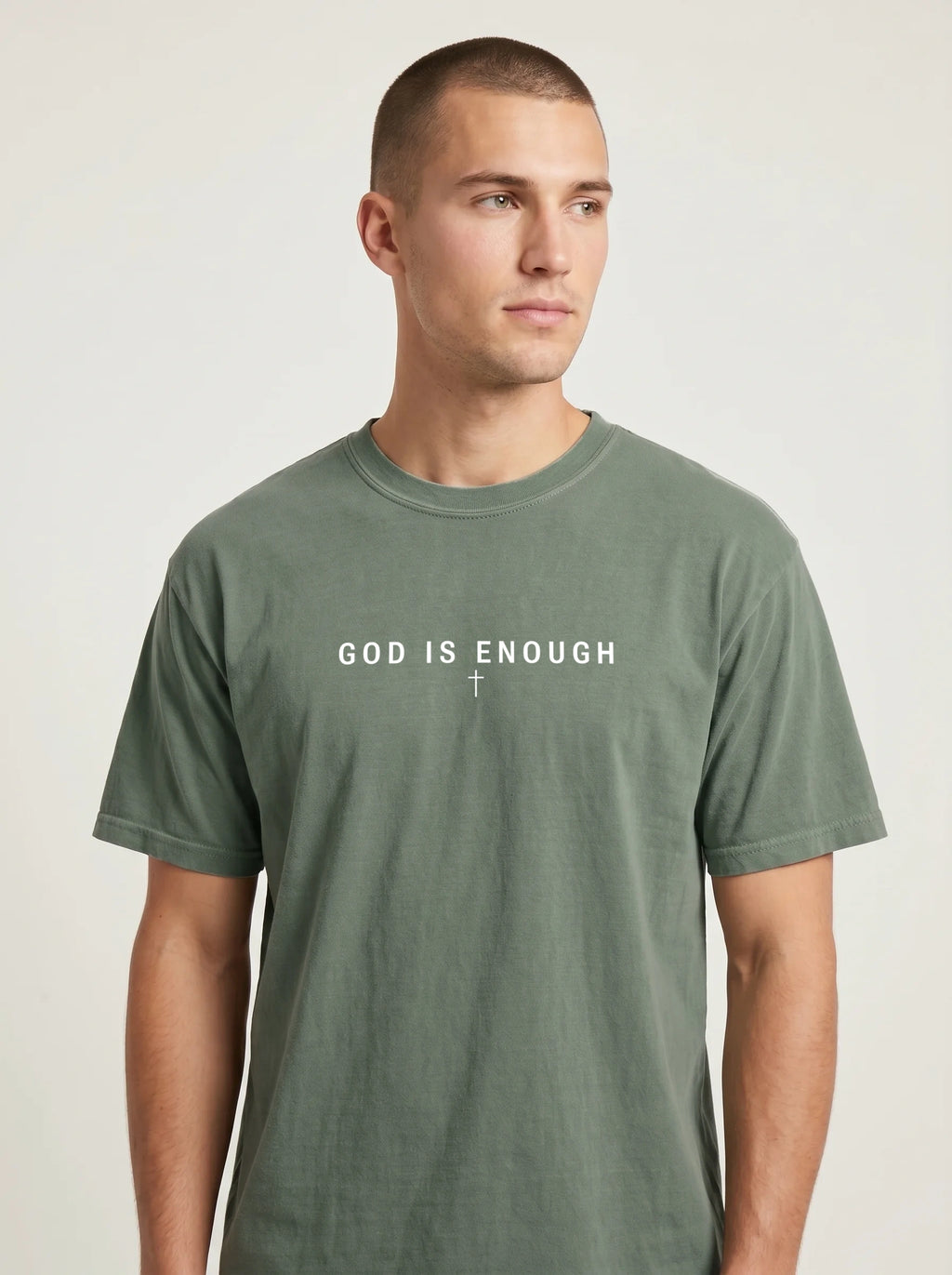 Person wearing a faded sage green T-shirt with the text “God is enough” and a small cross beneath it.