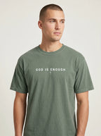 Person wearing a faded sage green T-shirt with the text “God is enough” and a small cross beneath it.