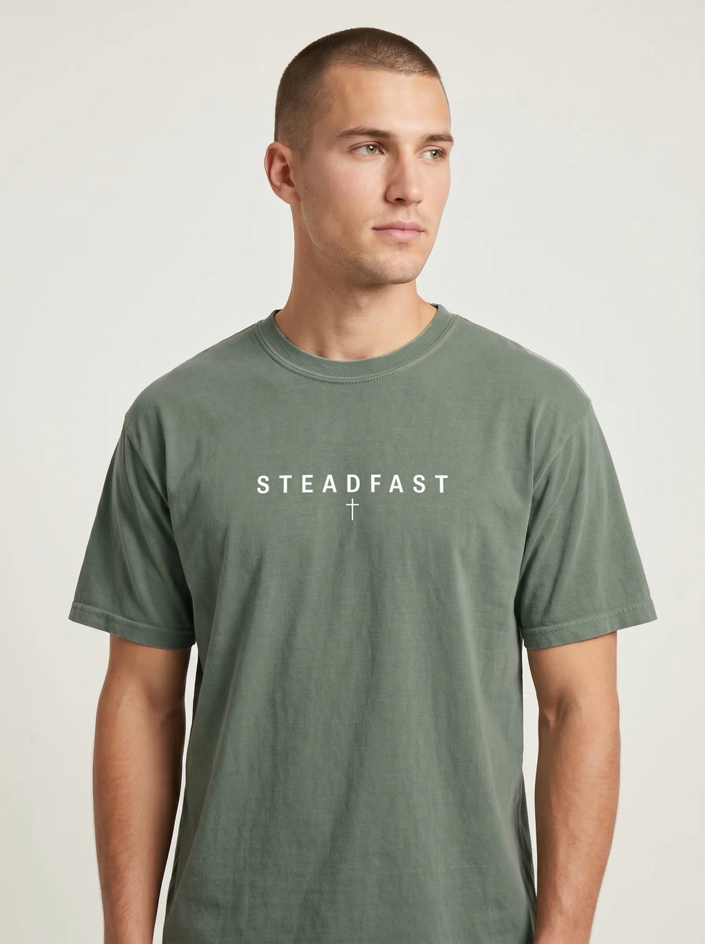 Person wearing a faded sage green T-shirt with the text “Steadfast” and a small cross beneath it.