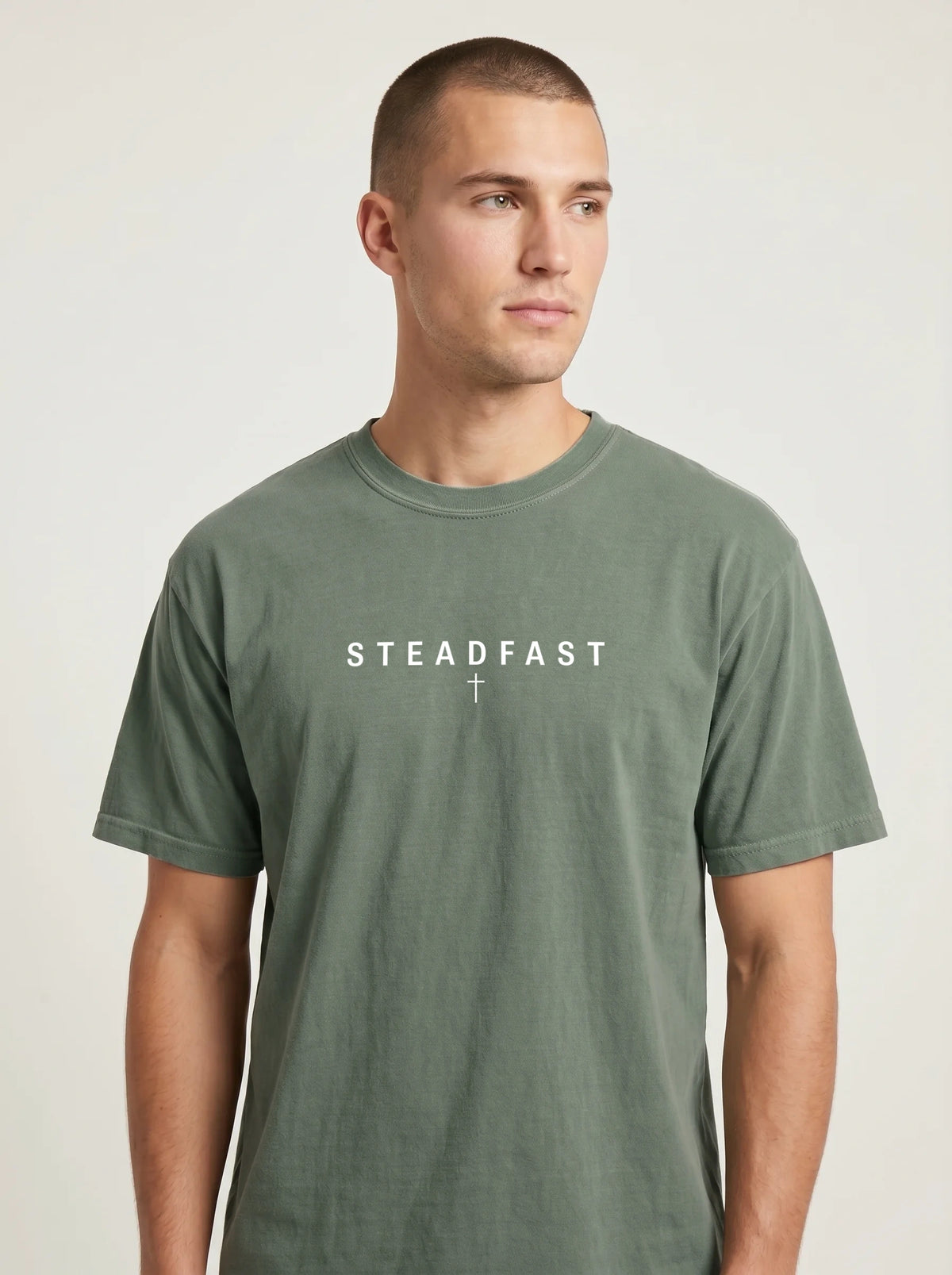 Person wearing a faded sage green T-shirt with the text “Steadfast” and a small cross beneath it.