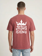 A man viewed from behind wearing a washed dusty rose t-shirt with a white graphic of a crown and the text 'JESUS IS KING' printed on the back, paired with light blue jeans against a light gray studio background.