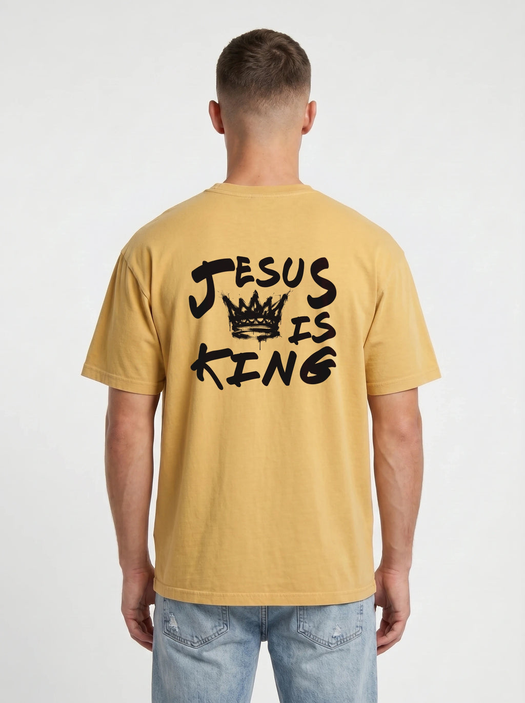 A man viewed from behind wearing a mustard yellow t-shirt with a black graphic print that reads 'JESUS IS KING' with a crown, paired with light blue jeans against a light gray background.