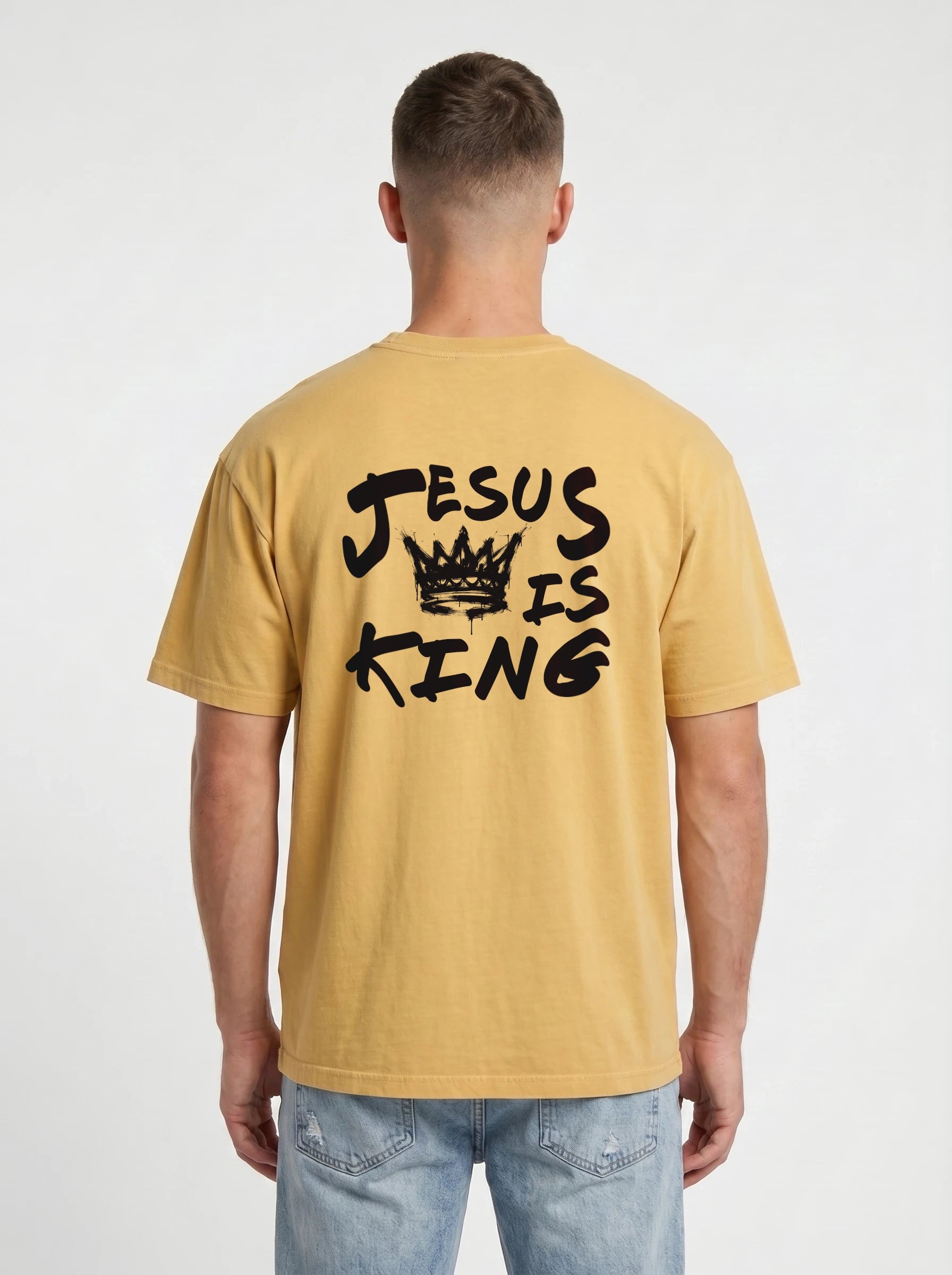 A man viewed from behind wearing a mustard yellow t-shirt with a black graphic print that reads 'JESUS IS KING' with a crown, paired with light blue jeans against a light gray background.