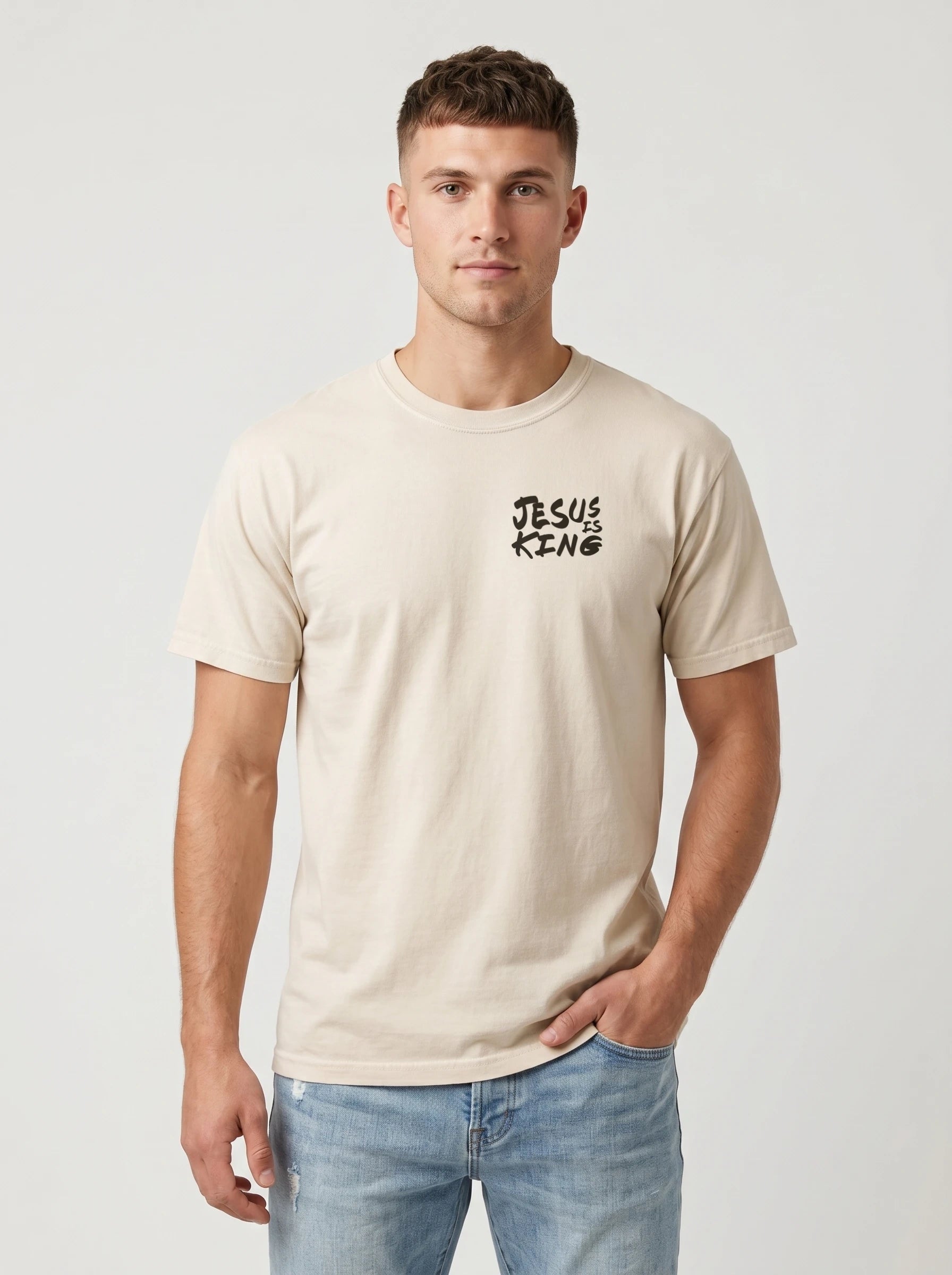 A man wearing a light beige crewneck t-shirt with a black 'JESUS IS KING' graphic on the left chest, paired with light blue jeans, against a light gray studio background.