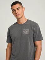 A man wearing a dark gray washed t-shirt with the words 'FAITH OVER FEAR' printed vertically in a light beige font on the left chest, photographed against a light gray studio background.