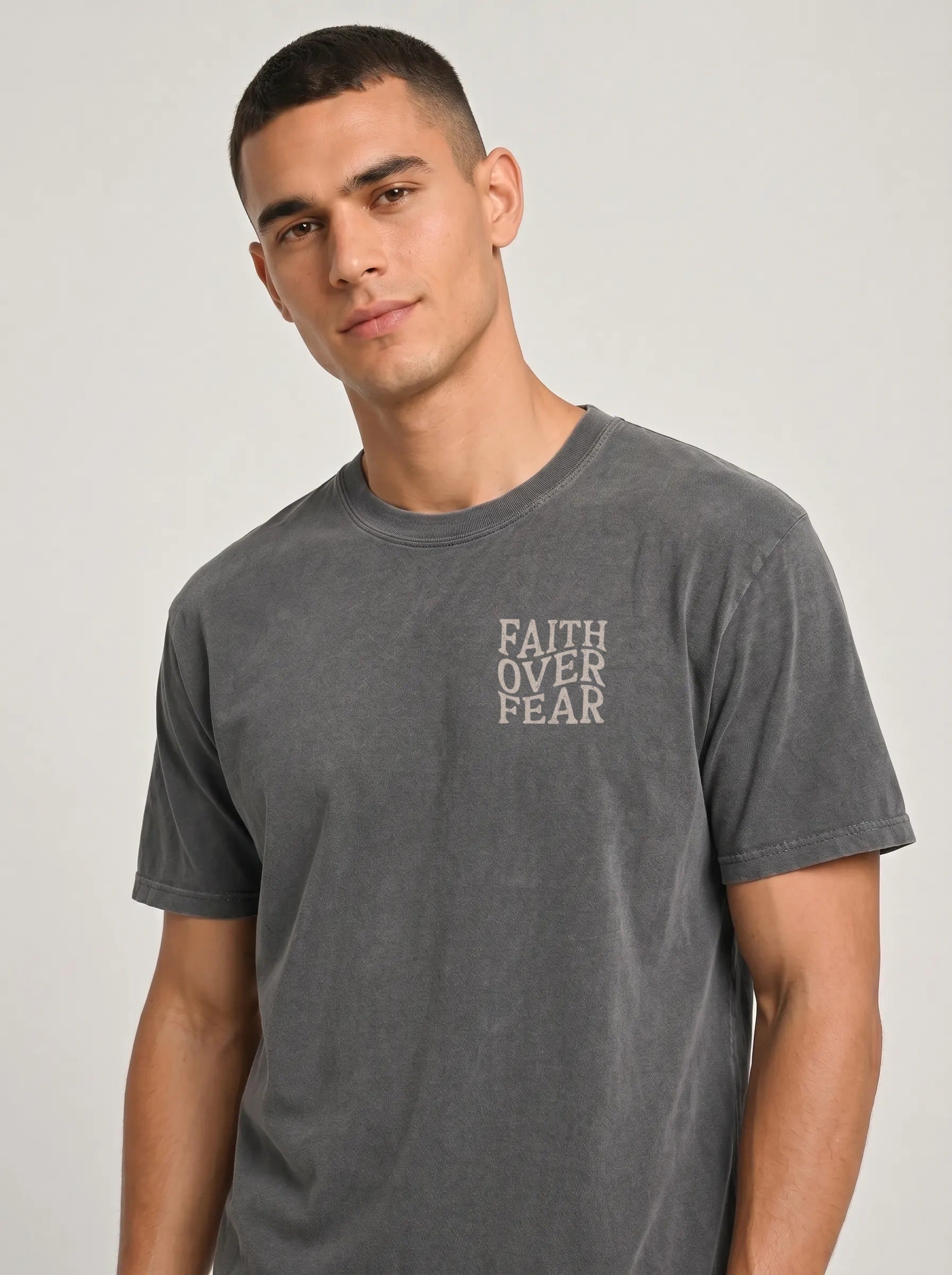 A man wearing a dark gray washed t-shirt with the words 'FAITH OVER FEAR' printed vertically in a light beige font on the left chest, photographed against a light gray studio background.