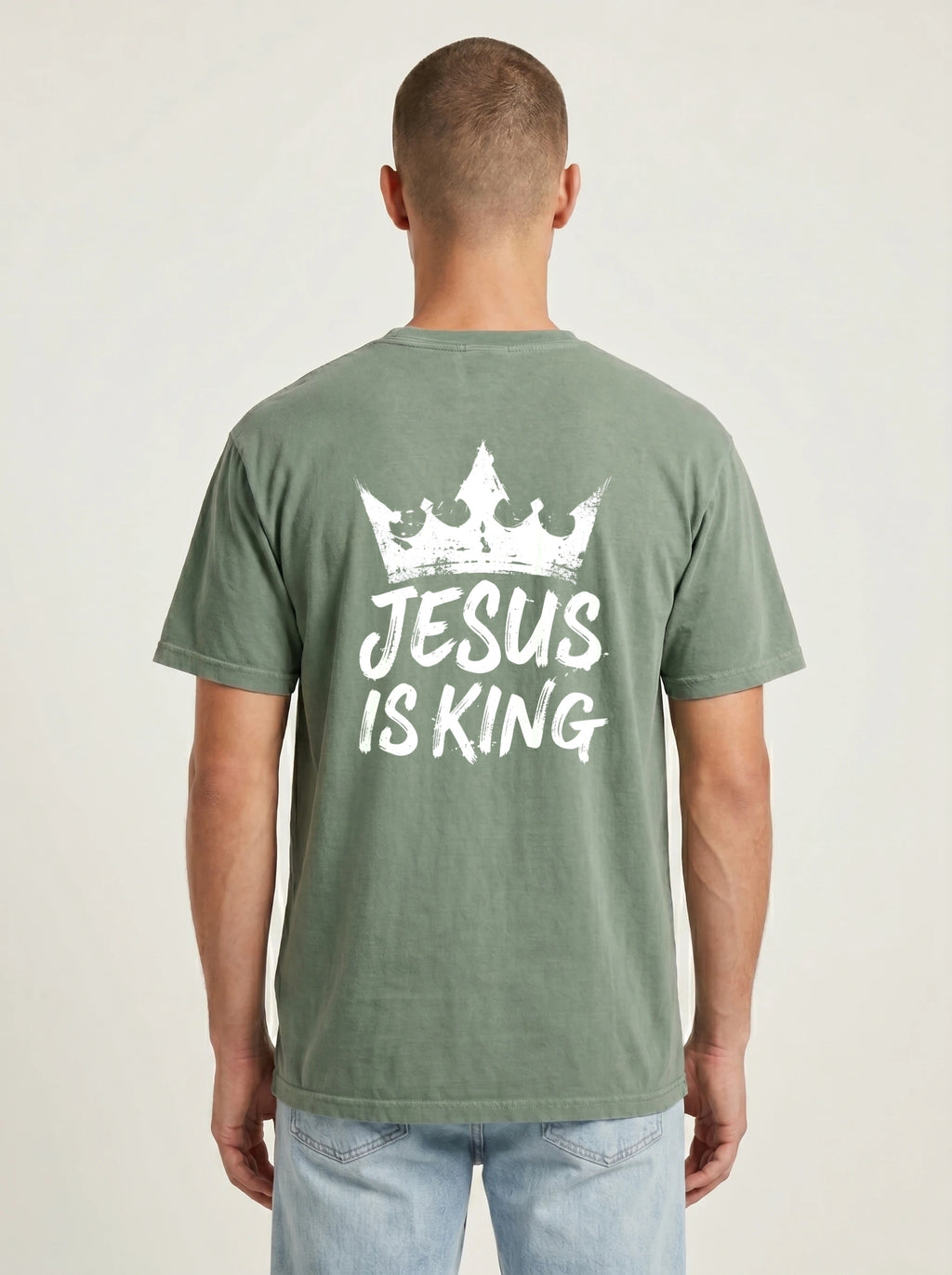 A man viewed from behind wearing a sage green t-shirt with a white graphic print of a crown and the text 'JESUS IS KING' on the back, paired with light blue jeans against a plain light gray background.