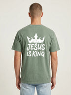 A man viewed from behind wearing a sage green t-shirt with a white graphic print of a crown and the text 'JESUS IS KING' on the back, paired with light blue jeans against a plain light gray background.