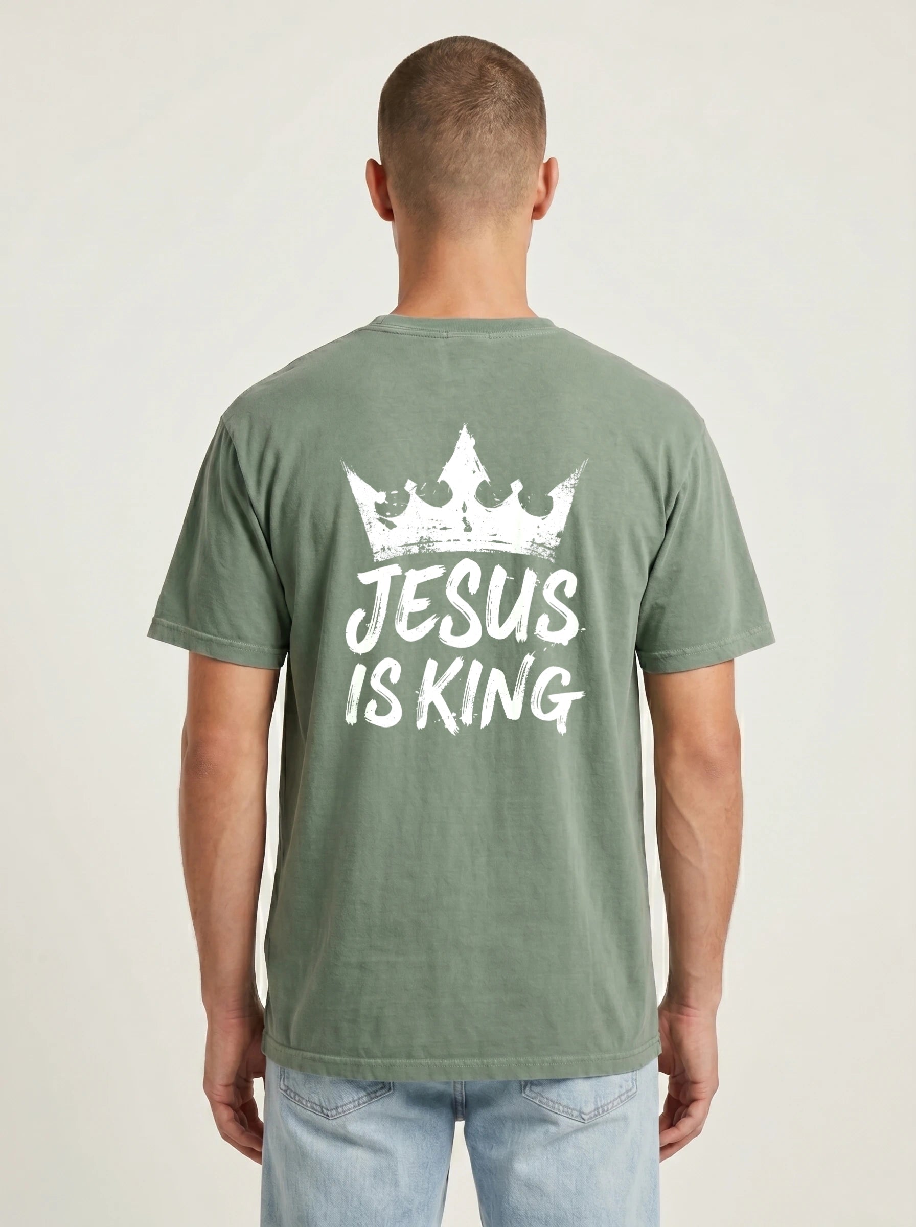 A man viewed from behind wearing a sage green t-shirt with a white graphic print of a crown and the text 'JESUS IS KING' on the back, paired with light blue jeans against a plain light gray background.