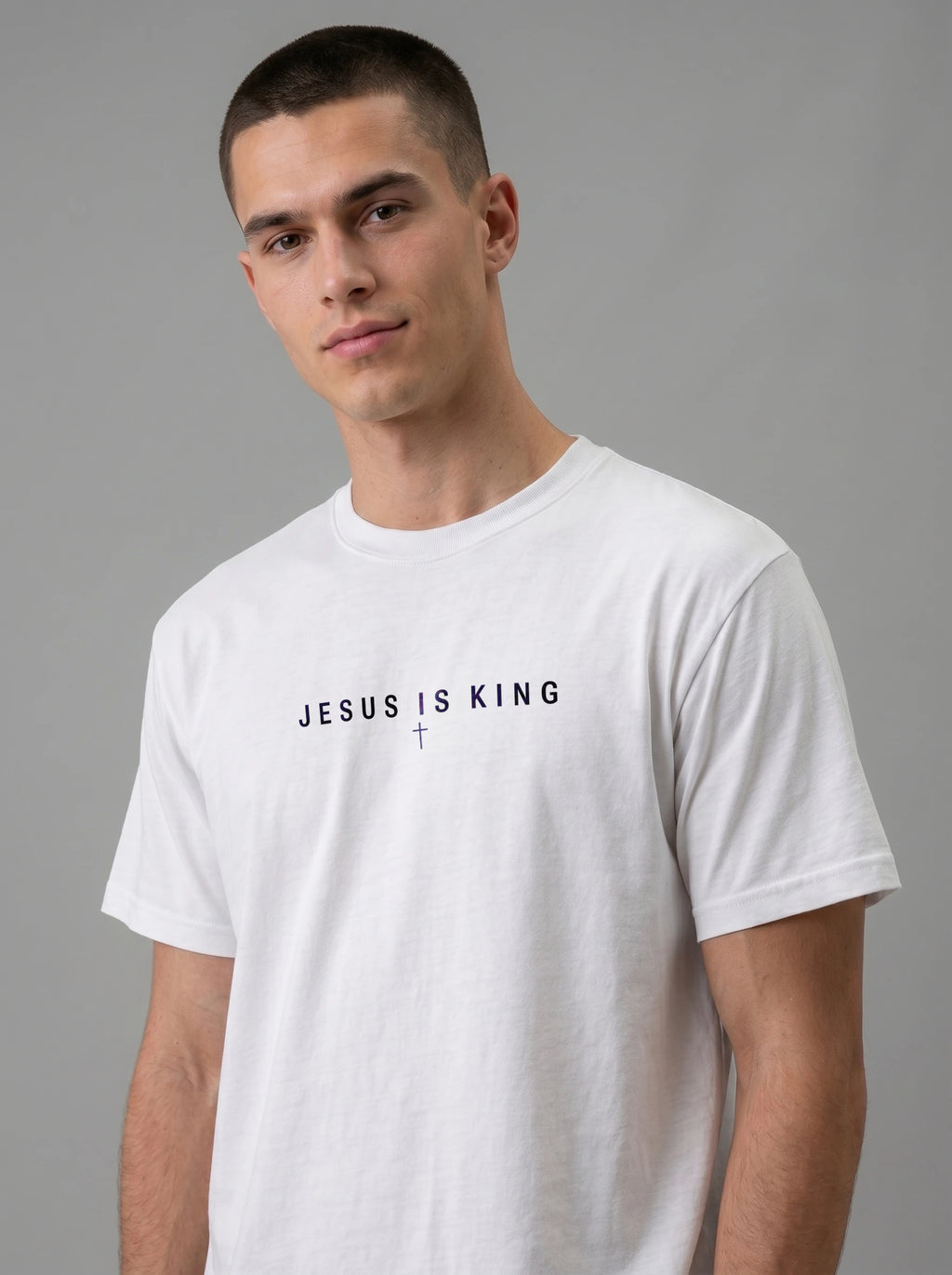 A man wearing a white crewneck t-shirt with the text 'JESUS IS KING' and a small cross printed in black on the chest, photographed against a solid gray studio background.