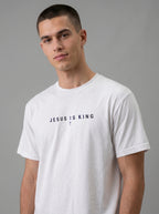 A man wearing a white crewneck t-shirt with the text 'JESUS IS KING' and a small cross printed in black on the chest, photographed against a solid gray studio background.