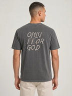 A man viewed from the back wearing a dark gray washed t-shirt with 'ONLY FEAR GOD' printed in a light beige gothic font on the back, paired with light beige pants, against a light gray studio background.