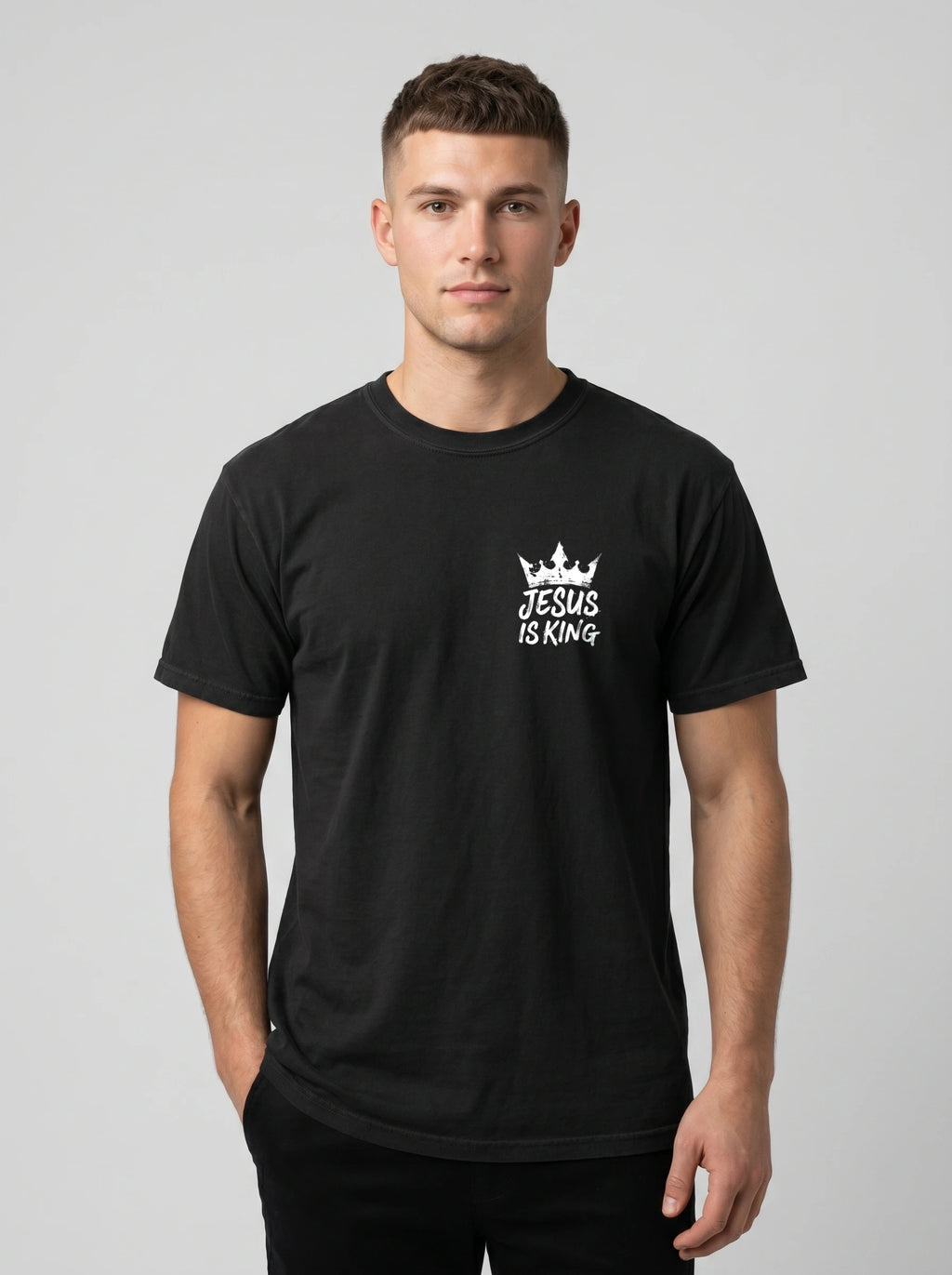 A man wearing a black short-sleeve crewneck t-shirt with a white graphic on the left chest that reads 'JESUS IS KING' beneath a small crown, photographed against a light gray studio background.