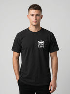 A man wearing a black short-sleeve crewneck t-shirt with a white graphic on the left chest that reads 'JESUS IS KING' beneath a small crown, photographed against a light gray studio background.