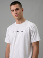Person wearing a white T-shirt with the text “Steadfast” and a small cross beneath it.