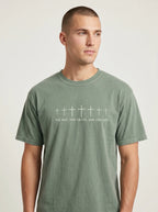 A man wearing a muted sage green crewneck t-shirt with the graphic of seven crosses in various sizes and the text 'THE WAY, THE TRUTH, AND THE LIFE', photographed against a neutral light gray background.