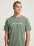 A man wearing a muted sage green crewneck t-shirt with the text 'STRENGTH' and the Bible verse 'ISAIAH 41:10' printed in white across the chest, set against a plain light gray background.