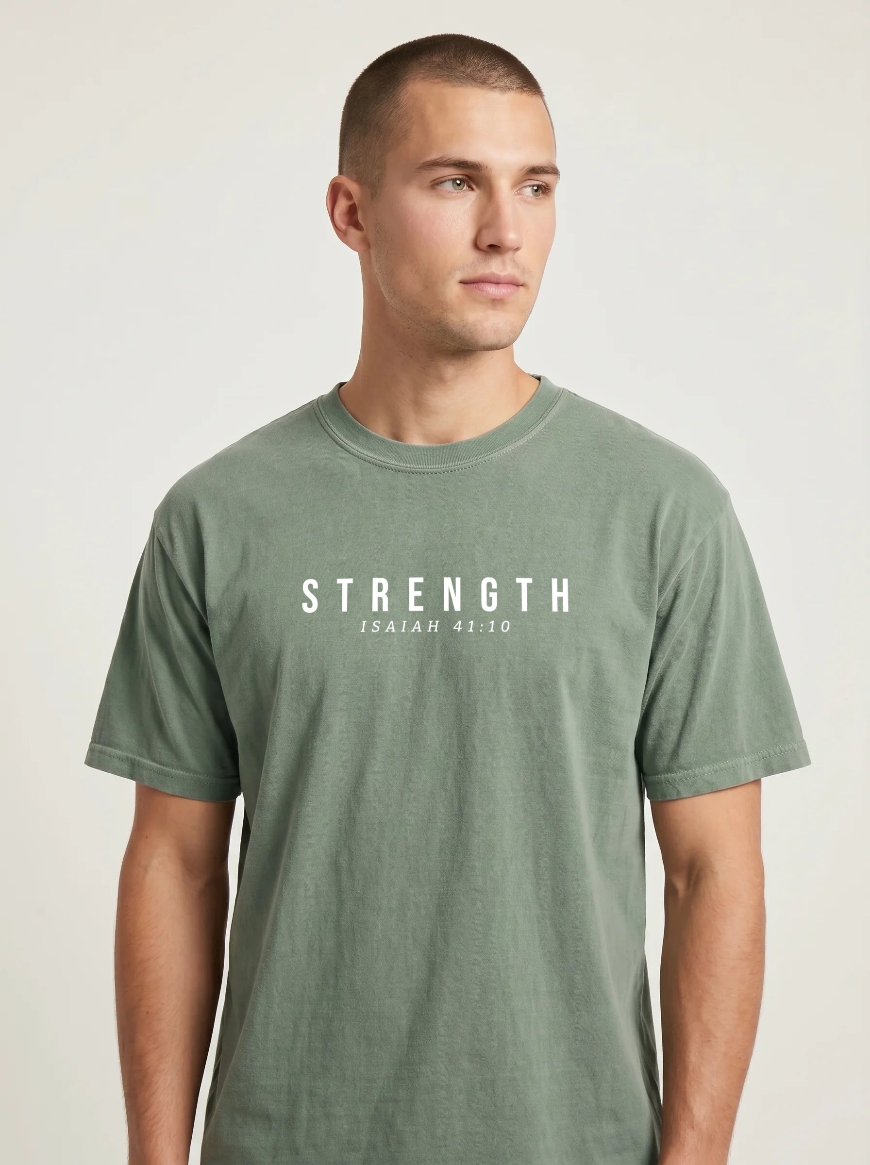A man wearing a muted sage green crewneck t-shirt with the text 'STRENGTH' and the Bible verse 'ISAIAH 41:10' printed in white across the chest, set against a plain light gray background.